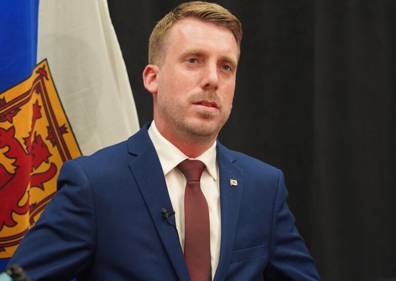 A man in a suit and tie stands in front of a flag.