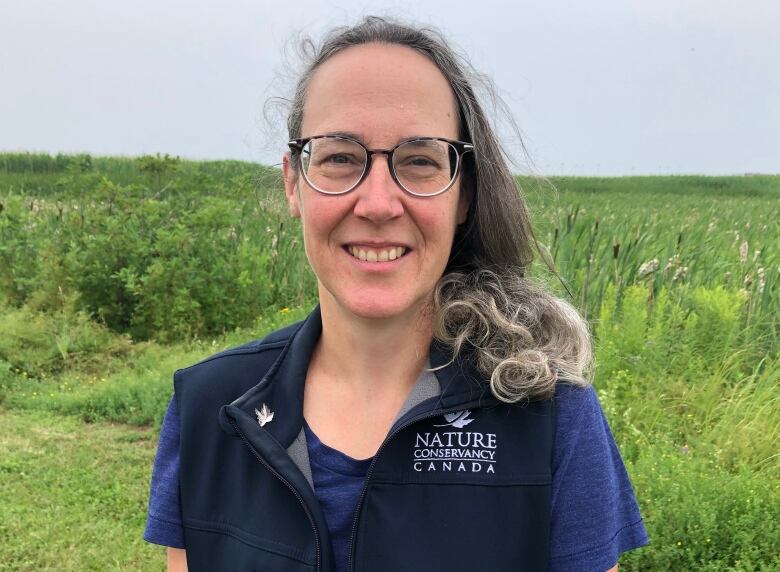 A smiling woman with long hair pulled over one shoulder. She is wearing a blue shirt and blue vest with a small logo on it that says Nature Conservancy of Canada