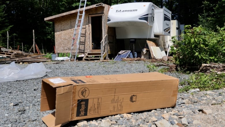 A long cardboard box sits on grey gravel in front of a large white camper. There is a wooden covered structure attached to the RV.