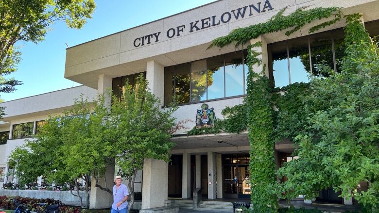 A man walks by a building with ivy on its pillars and the name 'City of Kelowna' over the entrance on a sunny day. The building is bordered by a flower bed with red pansies and a dark green lawn.