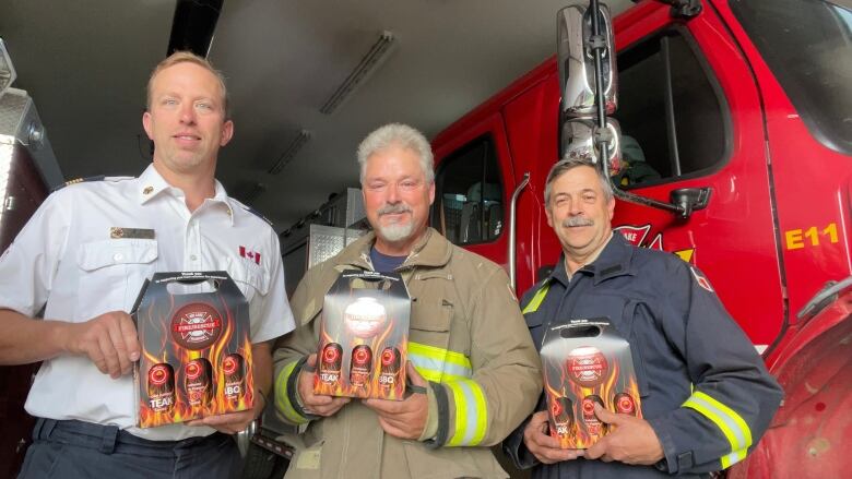 Three men stand holding three-packs of sauce. They are standing in front of a fire truck