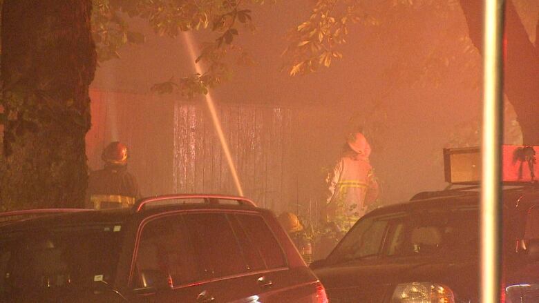 A firefighter in a beige uniform holds a fire hose blasting water onto the wooden siding at night. Grey smoke fills the air.