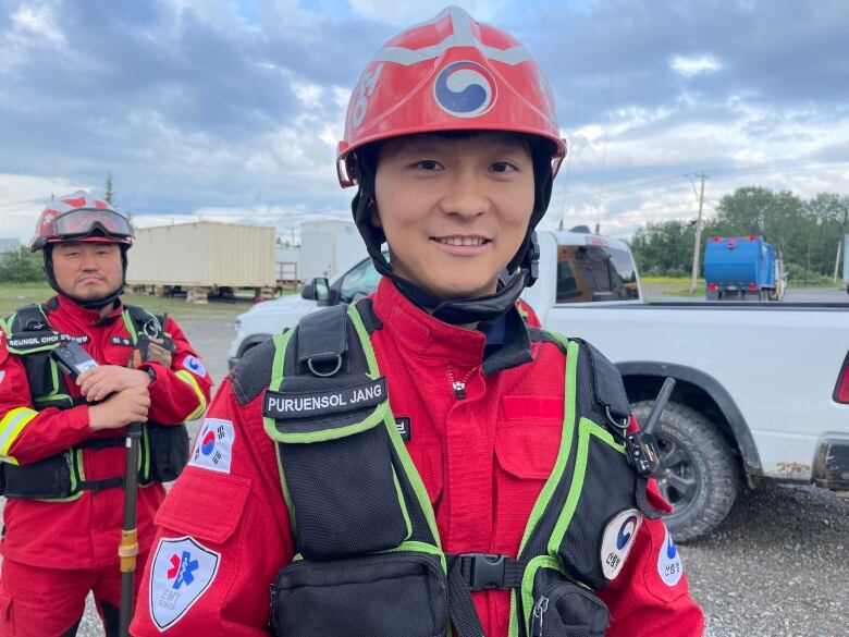 A young Korean man in a red helmet, red firefighting uniform and decked out in gear smiles into camera. 