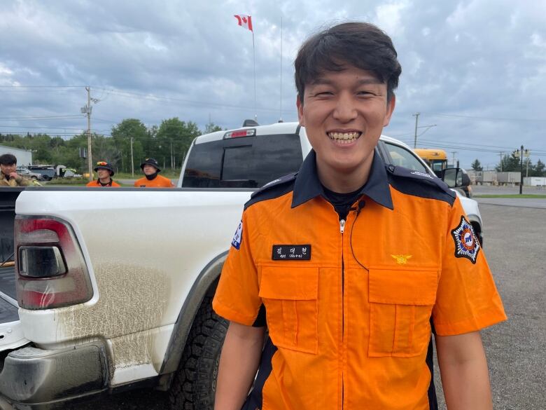 A Korean man in his thirties dressed in an orange firefighting uniform smiles to the camera. Behind him is a pickup truck and two other firefighters as well as a Canadian flag on a mast pole.