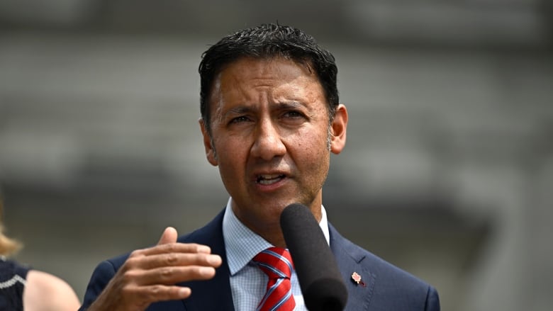 A man in a dark suit and red tie speaks at a microphone.