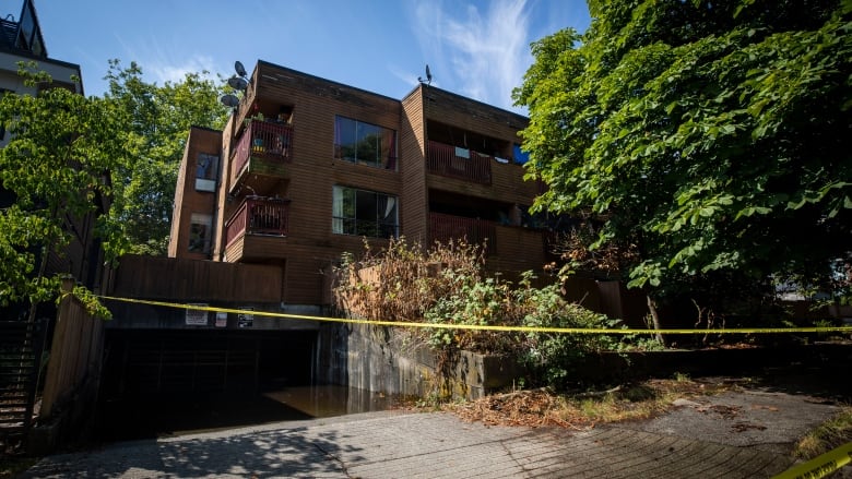 Caution tape is slung across the entrance to a lowrise brownstone apartment building surrounded by trees.