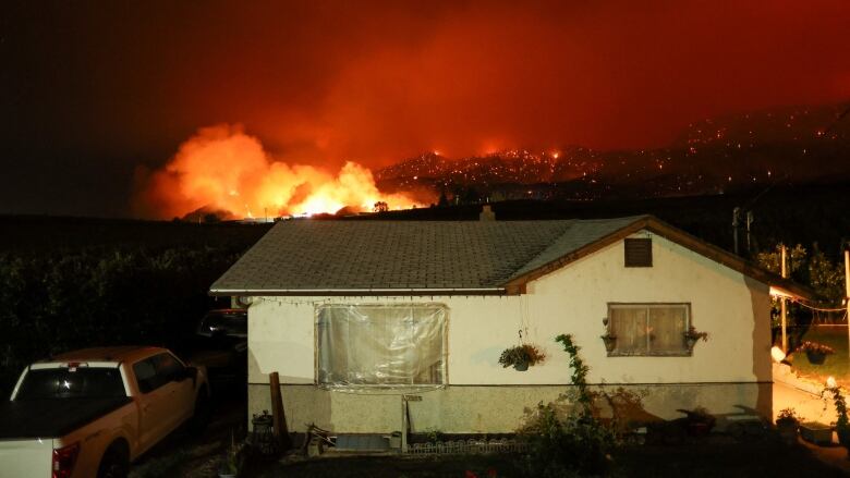 A fire burns prominently behind a small home with a pickup truck next to it.