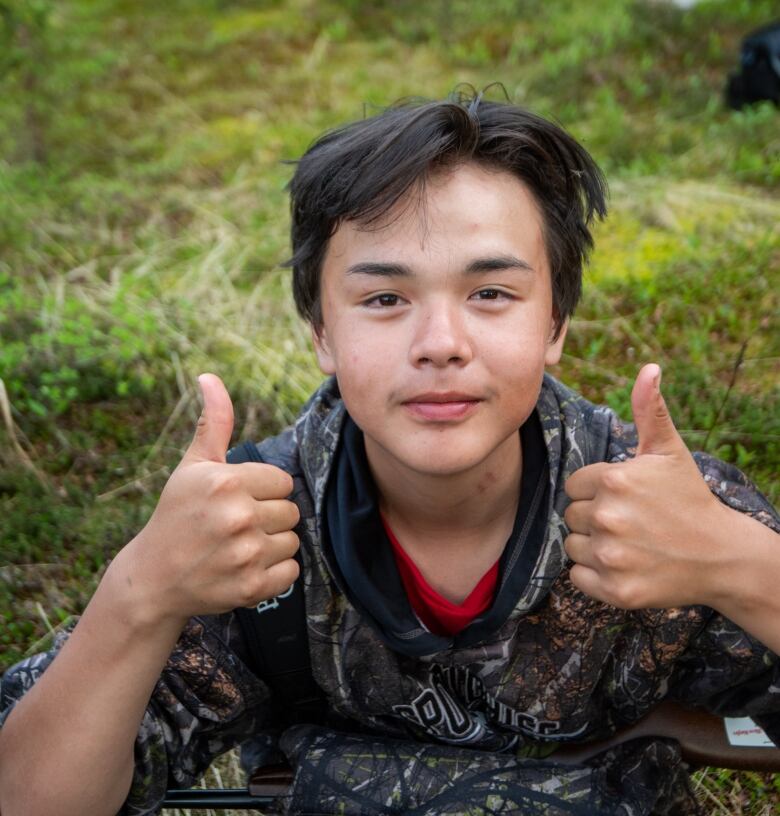 A boy holds two thumbs up while sitting on grass. 