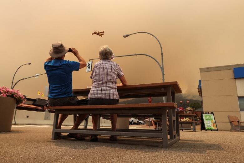 Two seniors sit at a picnic table and watch as a water bomber flies through the orange, smoky sky overhead. One of the seniors, a man wearing a blue shirt and a Tilly hat, holds binoculars to his eyes.