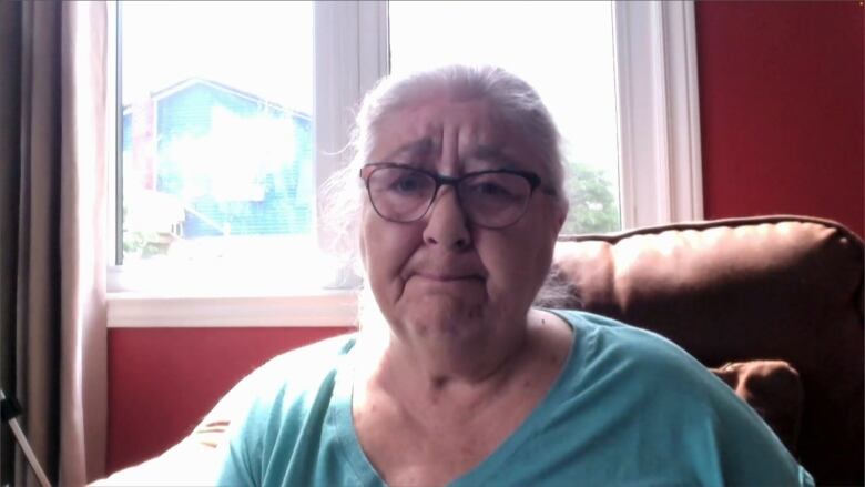 An older woman wearing a blue shirt sits in her home.