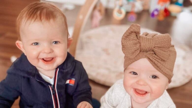 Two babies are smiling at the camera. They are twins. One is a boy and one is a girl. The girl wears a beige bow on her head. 