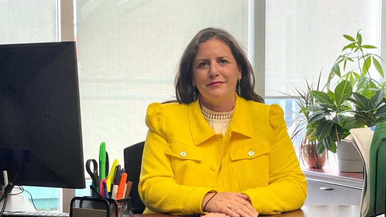 A woman with long brown hair and a yellow blazer sits at her desk. 