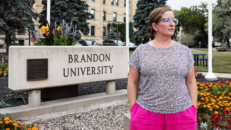 A scientist wearing glasses, a patterned shirt and pink pants looks on while standing in a front of a concrete Brandon University sign surrounded by red and orange flowers, with a three-and-a-half storey brick building in the background. 