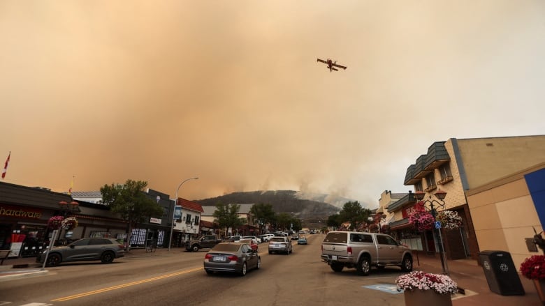 A water bomber soars through smokey skies while below a line of cars snakes through a small town street where baskets of flowers hang from lamposts and business appears to be continuing as usual. 