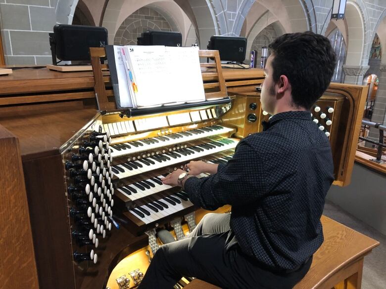 A man, seen from behind, on a bench playing an organ with four tiers of keyboards.