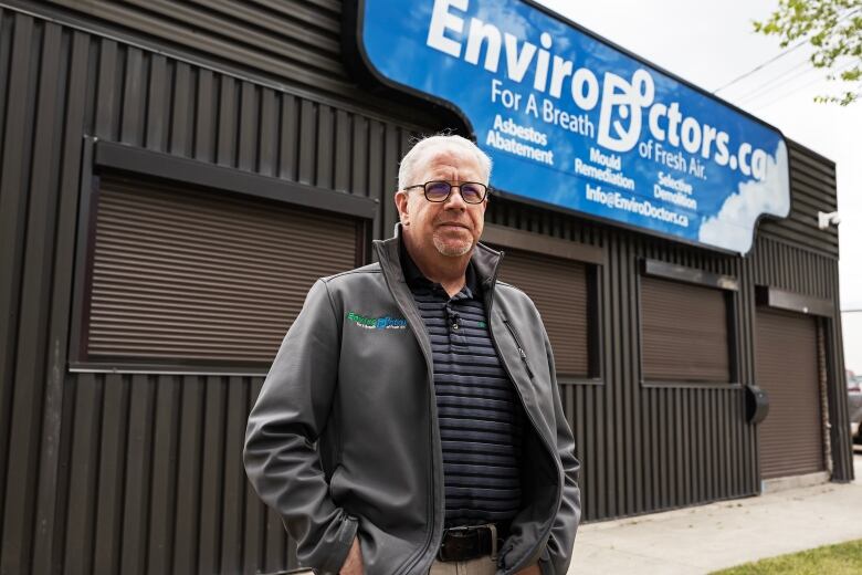 A man wearing a grey jacket, striped golf shirt and glasses stands in front of a business with a sign on that reads EnviroDoctors.