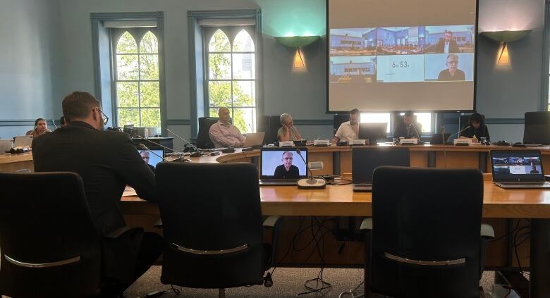 The back of a man at a round committee table with a screen showing a virtual meeting hanging in the background.