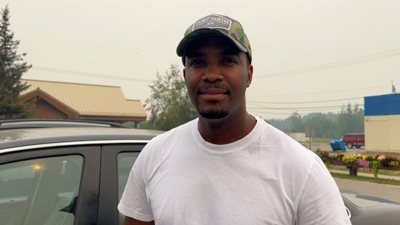 A man in a ballcap stands beside a car on a town street.