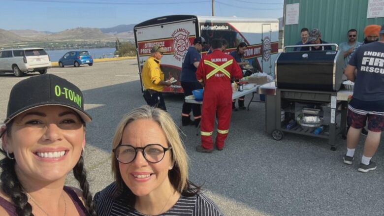 Two women pose with their heads together, photographed from the neck up in a 'selfie' style photo. The woman on the left has a big smile, two dark braids and is wearing a black hat that says O-Town in yellow font. To her right is a blond woman wearing black-framed glasses and also smiling. Behind them are a barbeque and a line of firefighters forming to get a meal.