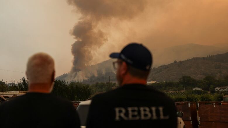 Two men watch as a wildfire on the other side of a large hill sends plumes of smoke into the air.