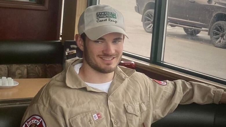 A young man is sitting in a diner wearing a brown collared shirt with a company emblem on the shoulder. He's smiling and looking sideways, with one arm over the top of the diner chair.