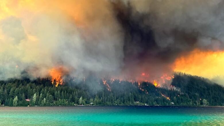 Flames rise from a forested hilltop next to a picturesque lake.