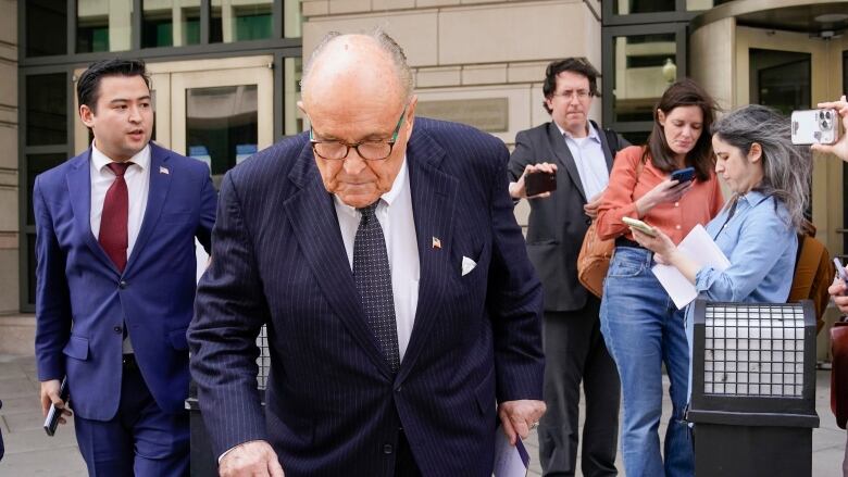 A balding man in a suit and tie is shown on the sidewalk outside a building.