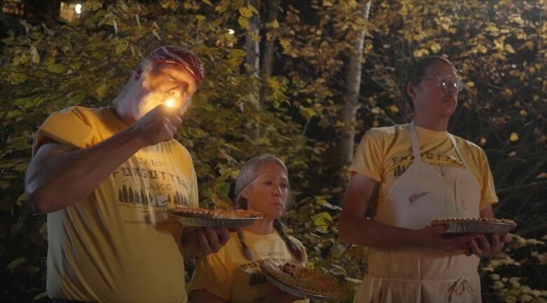 A still from a film shows three people in yellow shirts standing in the woods, each holding a pie.