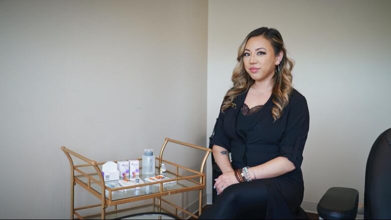 A woman sits next to a gold cart with syringes and bottles on it. 