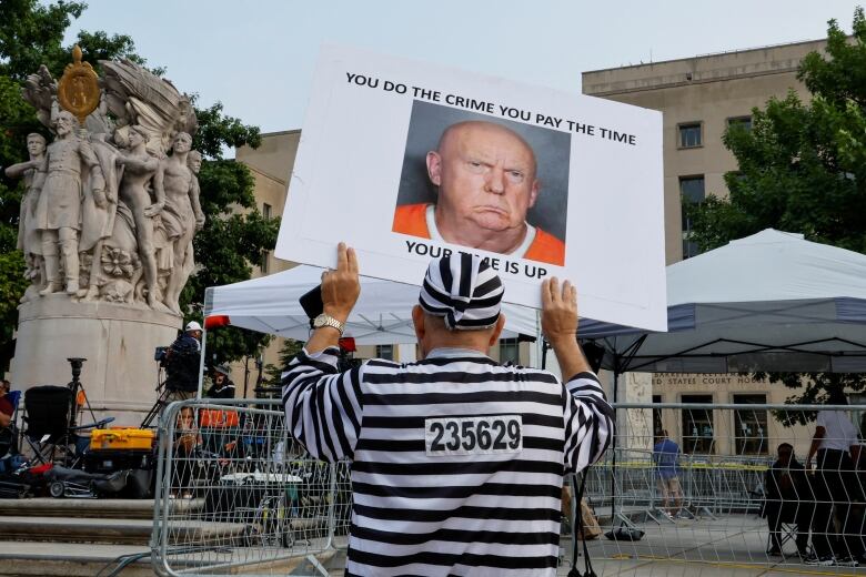 A demonstrator dressed in a prison-like uniform holds up a sign outside of metal barricades erected around a building. The sign reads: 