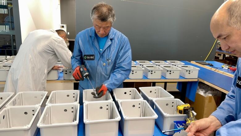 Three workers in lab coats use screwdrivers to assemble equipment.