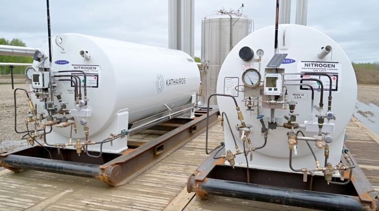 Two large white tanks sit on the ground at an oil and gas well site.