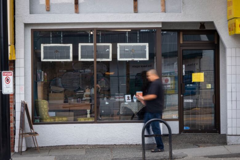 The exterior of the store is pictured. It is painted white with large windows trimmed in black paint. A man is passing by the windows and is blurred in the image with the store front in sharp focus.