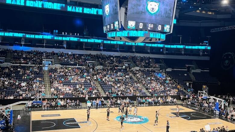 A basketball court is seen in a wide shot. Players are on the floor and fans in the stands.