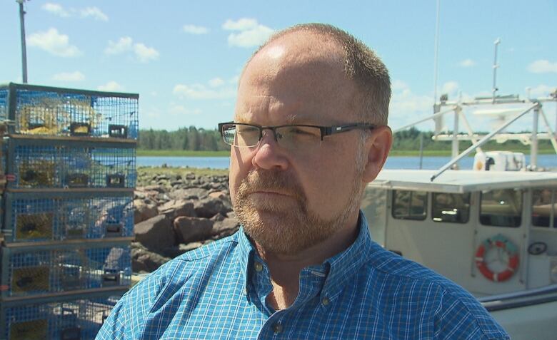 An unsmiling man wearing a blue plaid shirt and standing in front of a pile of lobster traps and a fishing boat.