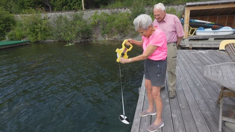 An elderly man and woman, both wearing khakis and pink shirts, stand at the edge of a dock looking down at lake water. The woman holds a tape measure with a weight on the ends to gauge the water's depth.