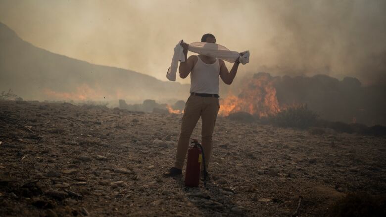 A man wearing a white tank top and long pants wraps a length of material around his face as he stands in a burning, smoky field with a fire extinguisher at his feet. Eye health experts are concerned that as wildfires become a more common phenomenon, we aren't studying the long-term impacts thesmoke could have on our eyes.