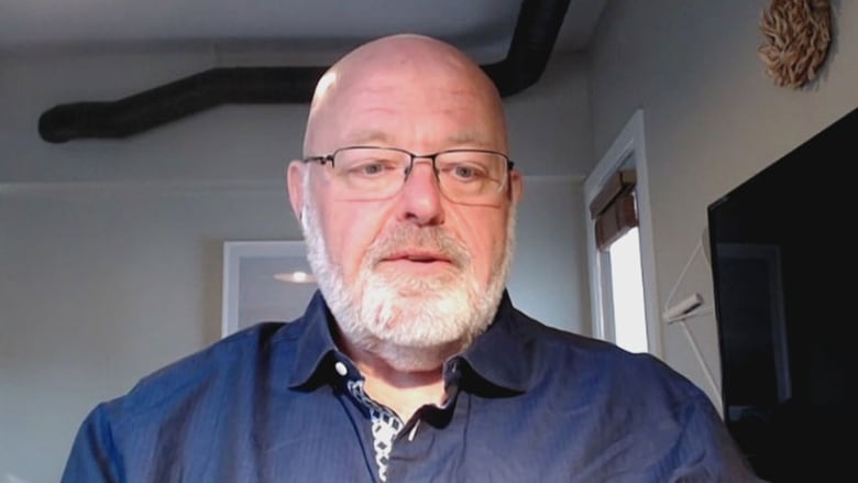 A bald man with a grey beard and glasses, wearing a blue collared shirt.