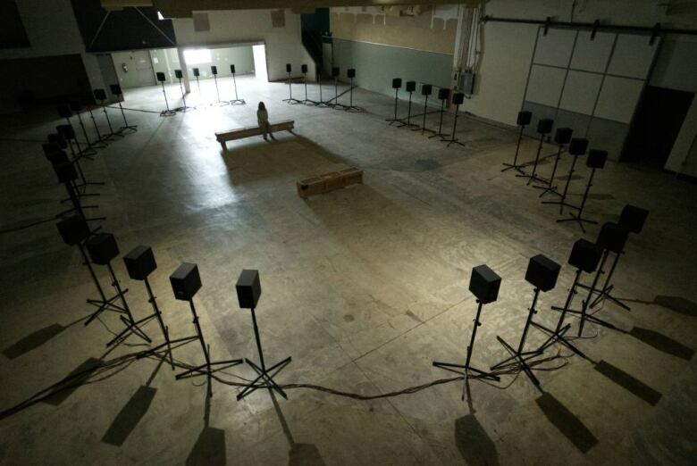 Shot from above, a view of a shadowy warehouse filled with a circle of 40 boxy speakers. A lone figure sits on a bench in the centre of the room.