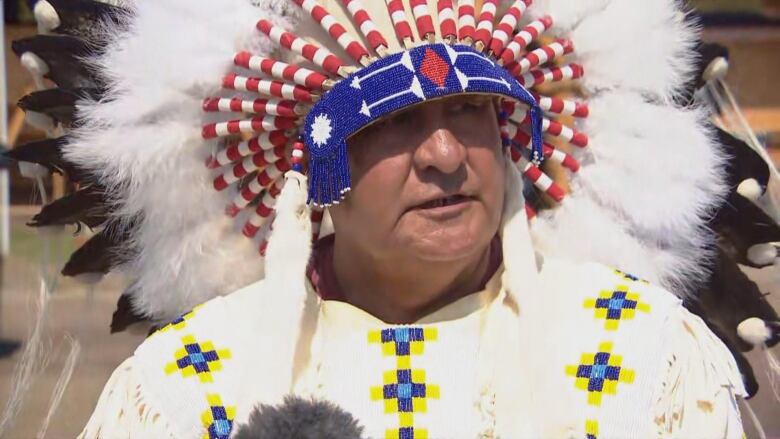 A Cree chief can be seen wearing his headdress, he is also wearing a cape in white, yellow and blue. 