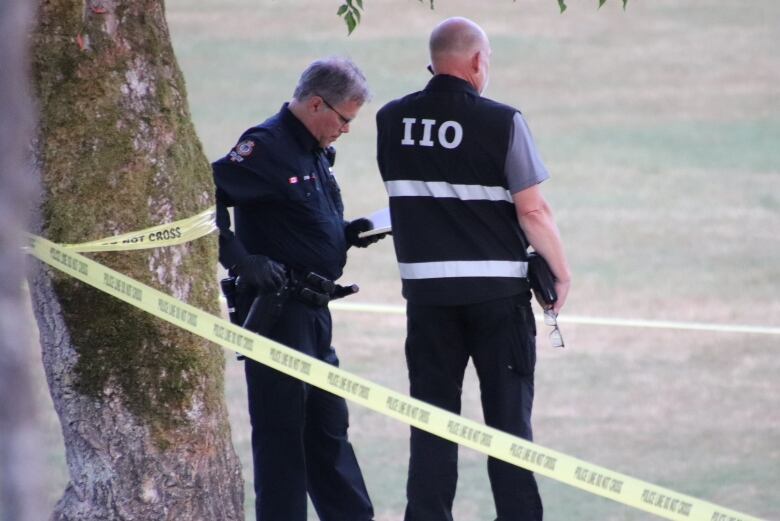 A man wearing a vest that reads 'IIO' is seen talking to a man wearing a police uniform behind police tape.