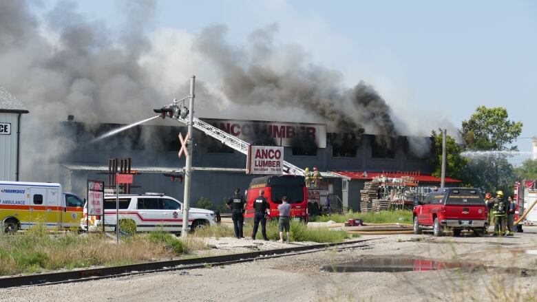 Emergency vehicles surround a lumber yard. A ladder used to spray water is pointed at the building as dark smoke billows from the windows.