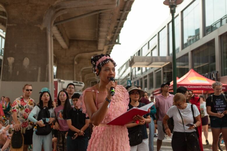 A person dressed in all pink speaks, holding a microphone and book, in front of a crowd. 