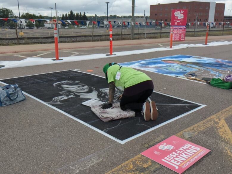 A person in a lime green tee-shirt, darker green baseball hat, black pants and dark sneakers crouches on a cushion in the middle of a chalk drawing of Elvis done on a street surface.