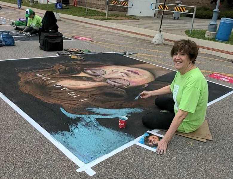 A woman with short brown hair wearing a lime green tee-shirt and black pants smiles up at the camera from where she is kneeling on part of her portrait of John Lennon, with the text 