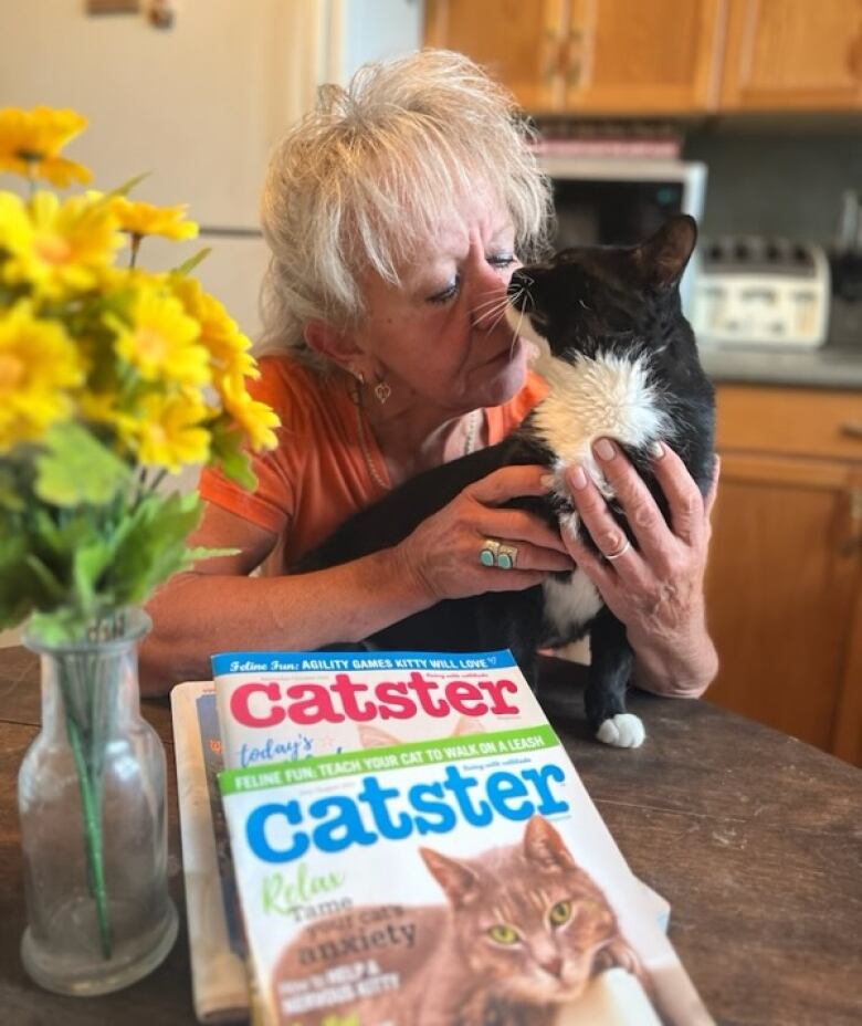 A woman is sitting in a kitchen holding a black tuxedo cat closely. There are several cat magazines on the table in front of her, and a vase of yellow flowers.