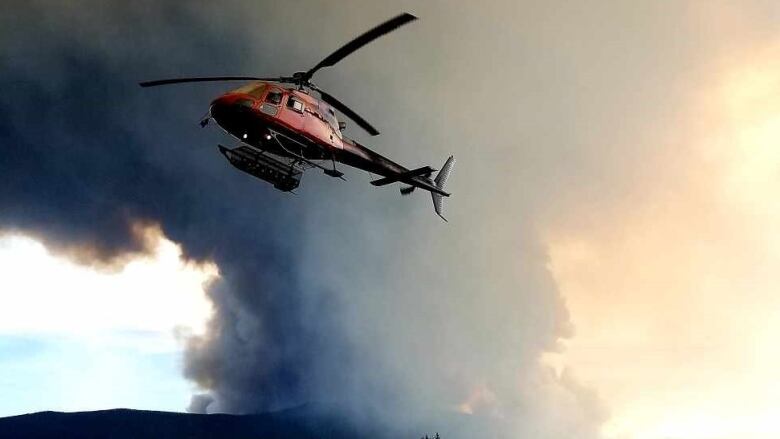 A helicopter is seen taking off with a big plume of wildfire smoke visible in the background.