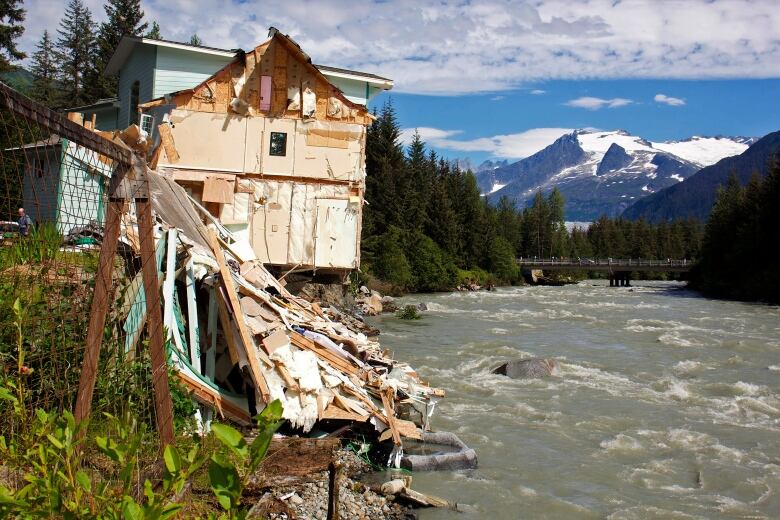 A partially-destroyed building is seen perched on the bank beside a fast-moving river.