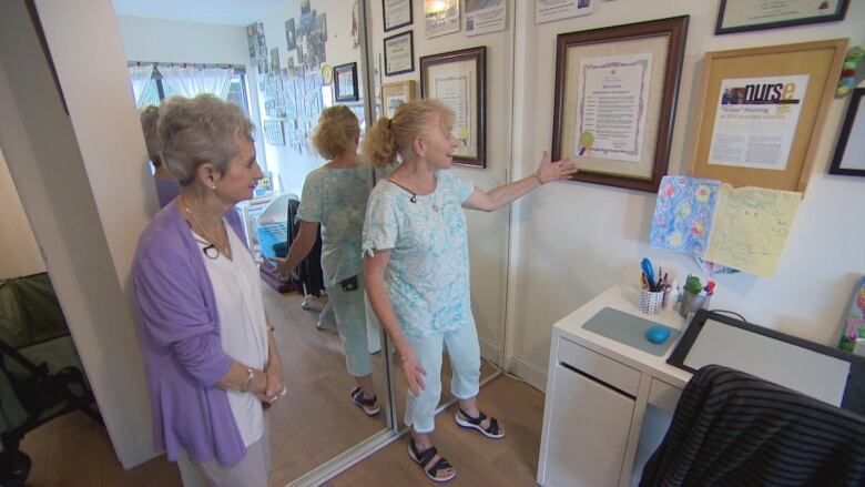 Two women point to awards and news clippings on a wall.