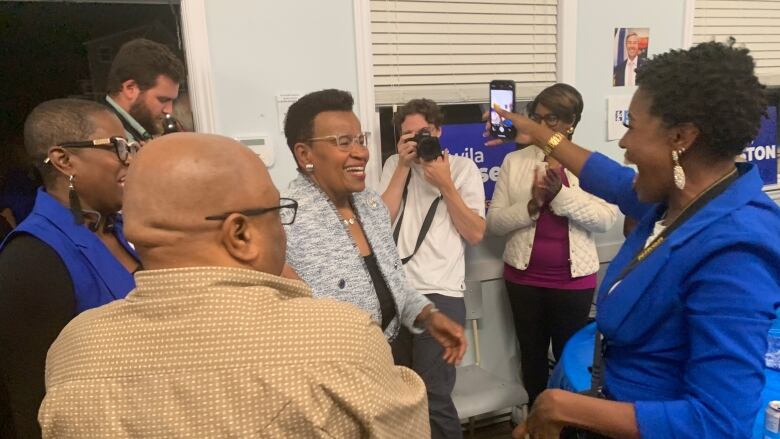 A woman wearing a light tweed blue blazer and glasses is smiling. She is surrounded by other people who are smiling.